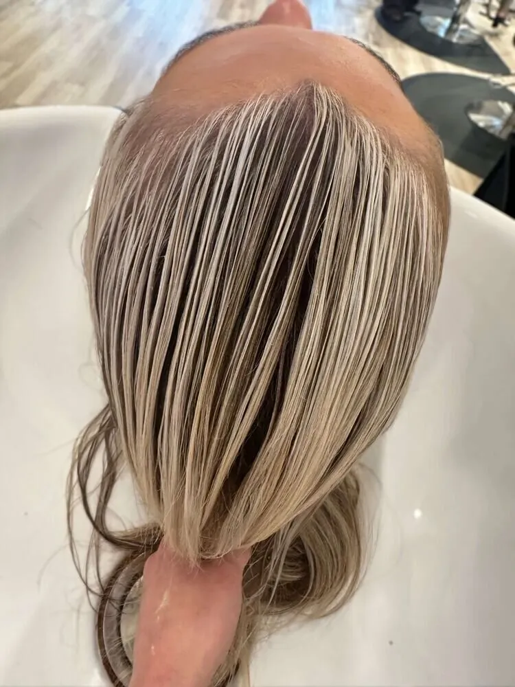 Client at a shampoo bowl with hair fanned out and rinsed, fine blonde highlights clearly separated through dark roots in an overhead salon view