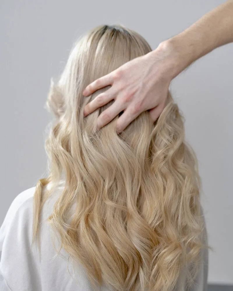 Stylist's hand gently lifting full, voluminous wavy blonde hair on a client seen from behind, against a clean light gray background