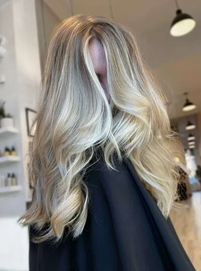 Long wavy hair with multi-tonal blonde color — bright blonde ends blending softly into a deeper root — draped over a black cape in a bright salon